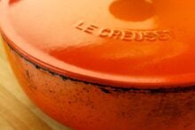 Close-up of an orange Le Creuset cast iron Dutch oven with a worn, slightly chipped edge, sitting on a wooden surface. The brand name "Le Creuset" is embossed on the lid.