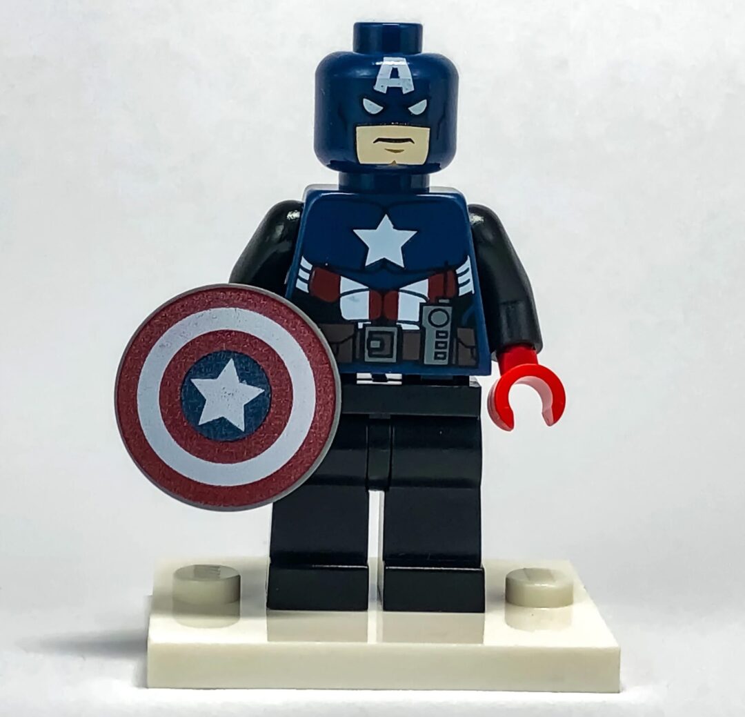 A LEGO figure dressed as a superhero with a blue helmet featuring an "A", a star emblem on the chest, and holding a red, white, and blue shield. The figure stands on a white base against a plain white background.