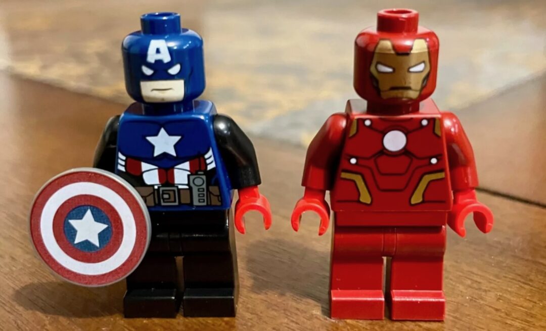 Two LEGO minifigures stand on a wooden surface. The left figure resembles a superhero with a blue suit and a shield with a star emblem. The right figure is in a red suit with a metallic mask resembling a tech-themed superhero.