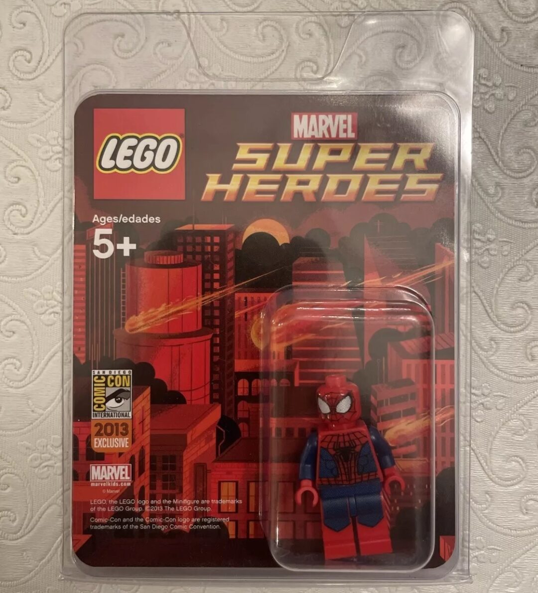 A LEGO Super Heroes set featuring a Spider-Man minifigure is sealed in its packaging. The background shows a cityscape with a red and orange design. The packaging includes Comic-Con 2013 exclusive branding.