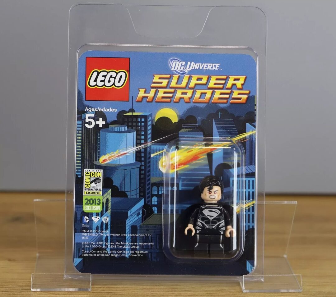 LEGO DC Universe Super Heroes set featuring a Superman minifigure in black suit, sealed in packaging. The package displays a comic-style cityscape with a streaking meteor. A label indicates San Diego Comic-Con 2013 exclusive.