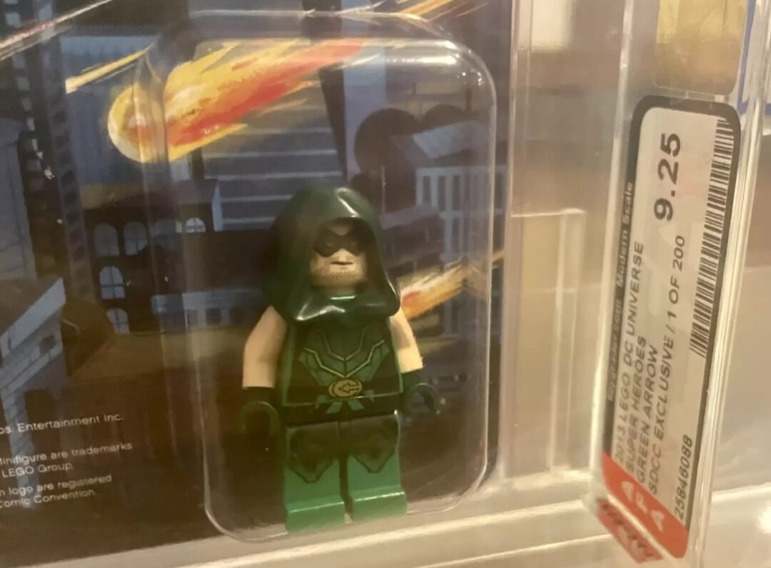 A collectible Green Arrow LEGO minifigure is displayed in a protective case. It features the character in a dark green hooded outfit. A label on the case shows a grading of 9.25.