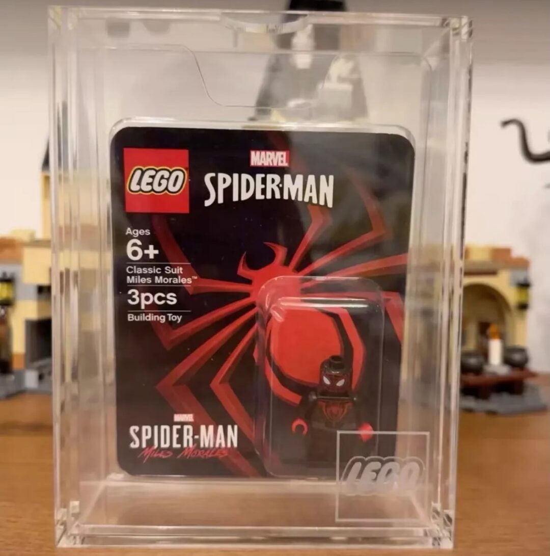 A LEGO Marvel Spider-Man classic suit Miles Morales minifigure in a protective display case. The packaging indicates it's for ages 6 and up and consists of 3 pieces. The background shows blurred LEGO structures.