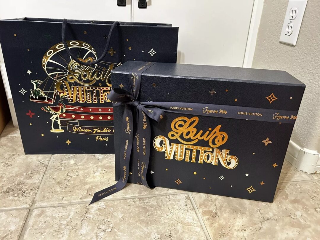 A large Louis Vuitton gift box with a ribbon sits on a tiled floor next to a matching shopping bag. Both are dark with gold and red accents, featuring the Louis Vuitton logo and decorative elements.