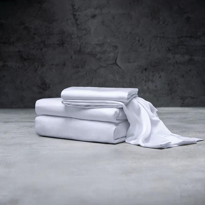 A neatly folded stack of white bed linens is placed on a smooth concrete floor with a textured, cracked wall in the background.