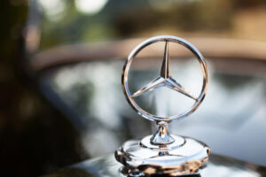 Close-up of a silver Mercedes-Benz hood ornament with a blurred background. The iconic three-pointed star emblem, often sought in high-end Mercedes-Benz auctions, is prominently displayed on the polished surface, suggesting luxury and elegance.