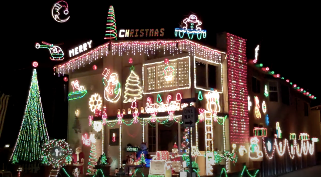 A house decorated with colorful Christmas lights, including a tree, snowman, candy canes, and Santa. The words "Merry Christmas" are illuminated. The lights cover the house's facade, creating a festive and bright display.