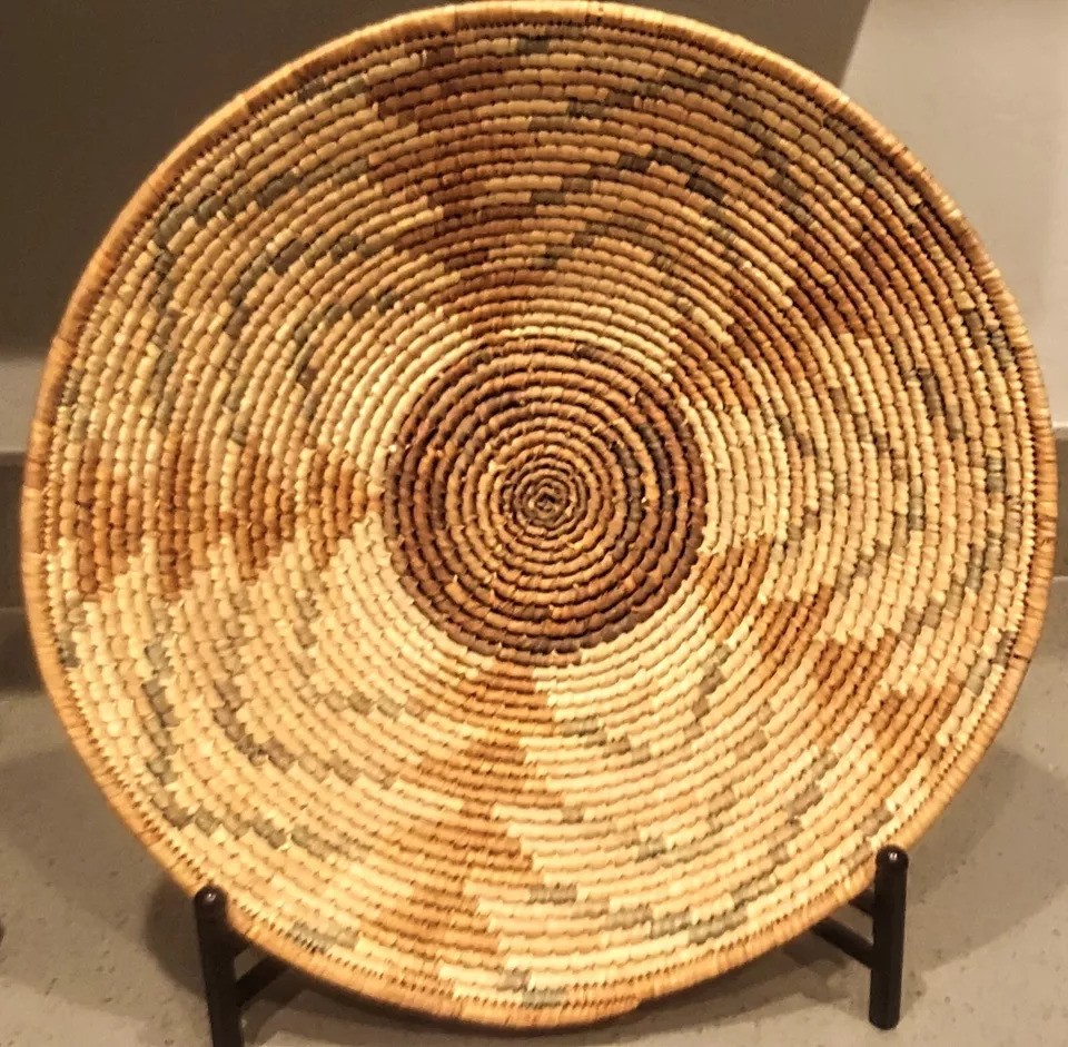 A round, woven basket with a spiral pattern of alternating light and dark natural fibers. It is displayed upright on a stand in a gallery setting.