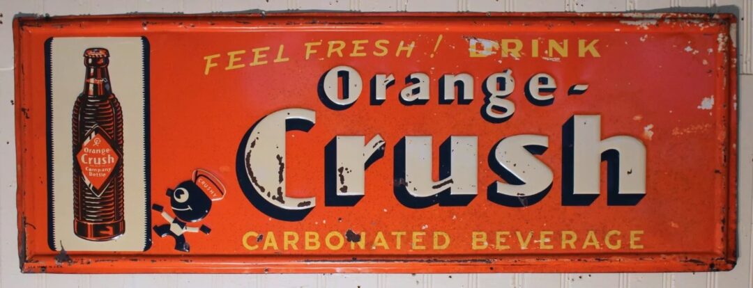 Vintage Orange Crush sign with a worn look. It features a bottle illustration and a small character. Text reads "Feel Fresh Drink Orange Crush Carbonated Beverage" on an orange background.