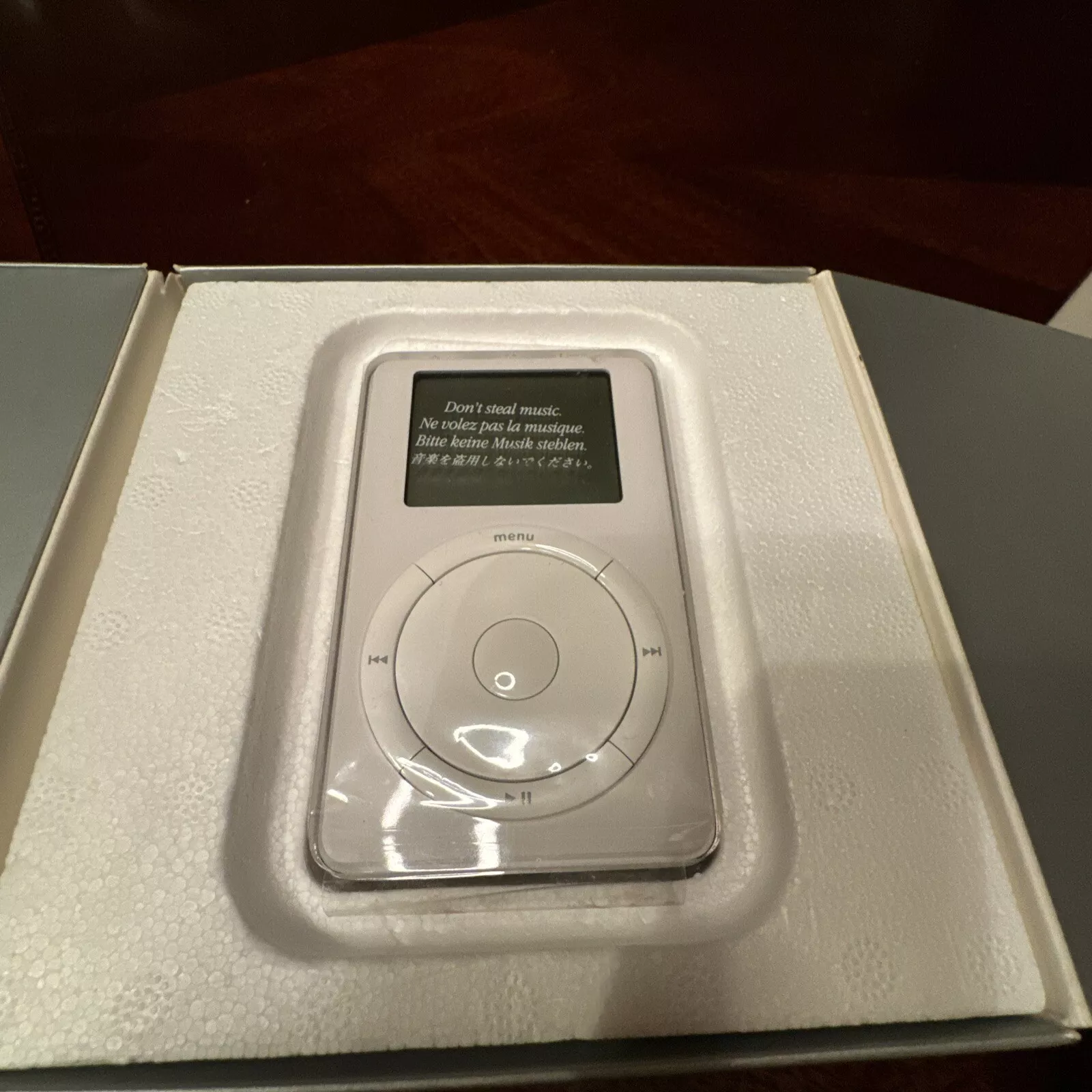 A classic iPod in its original packaging, surrounded by protective foam. The screen displays a message instructing not to steal music. The iPod has a circular control pad below the screen.