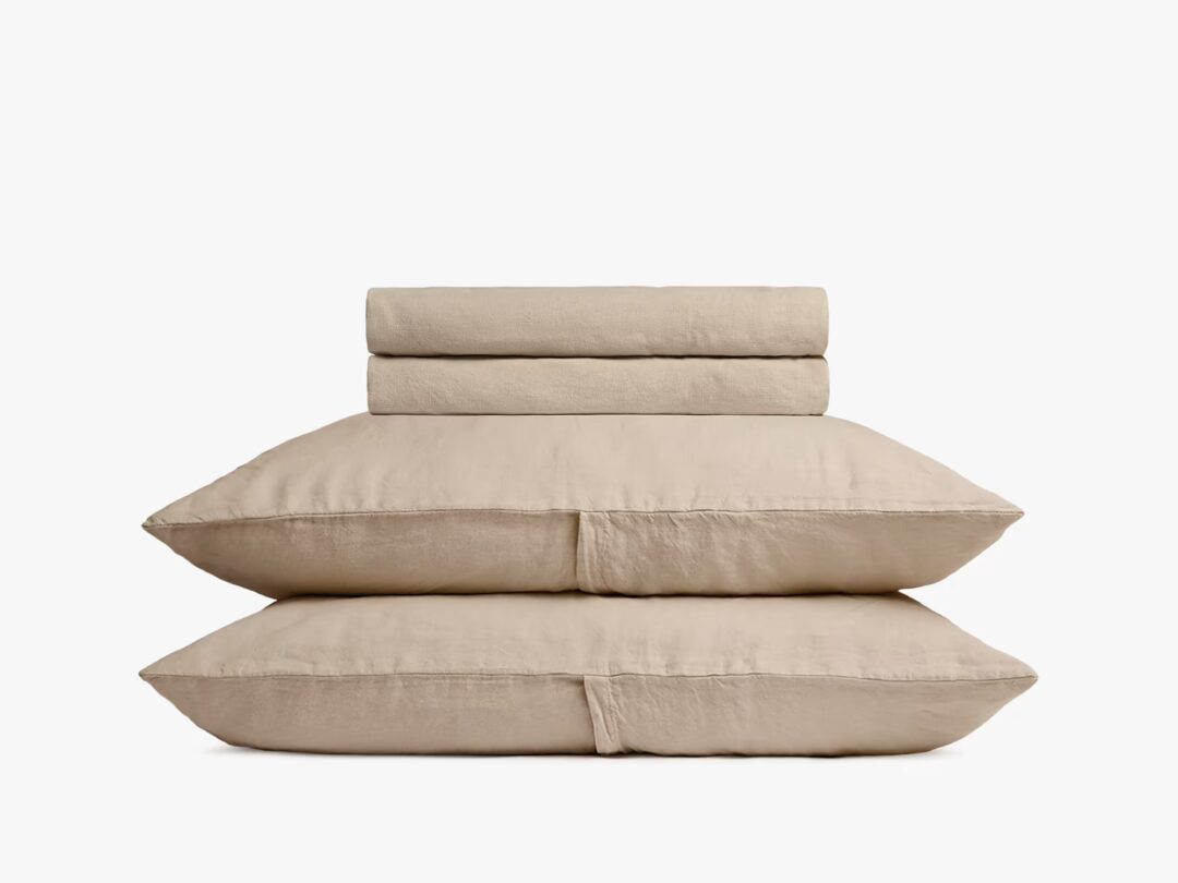 A neatly stacked set of beige bed linens, including two pillows, a folded duvet, and a folded sheet, against a plain white background.