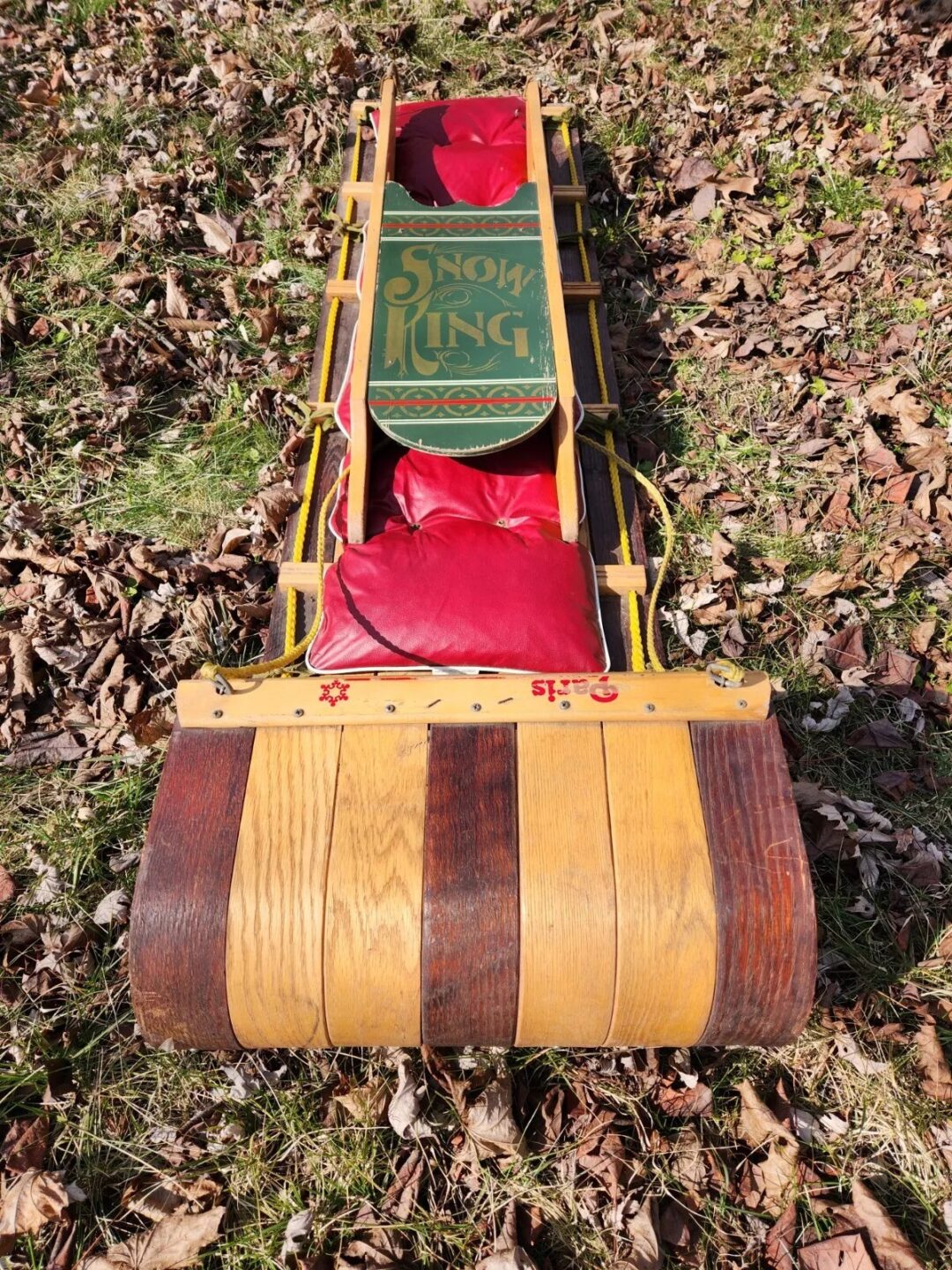 A vintage wooden sled with "Snow King" written on it, featuring red cushions. The sled rests on grass covered with brown fallen leaves.