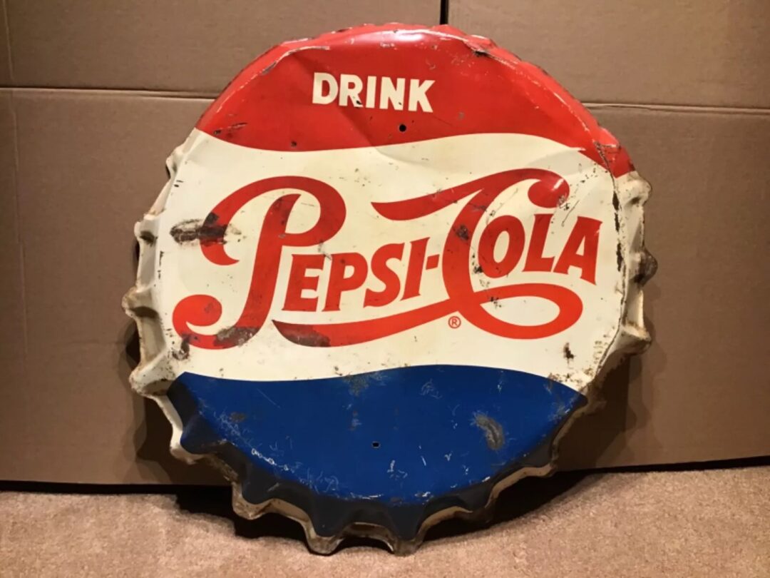 Vintage Pepsi-Cola sign shaped like a bottle cap with red, white, and blue sections. The sign is weathered and rusted, featuring the classic "Drink Pepsi-Cola" logo. It leans against a cardboard background.