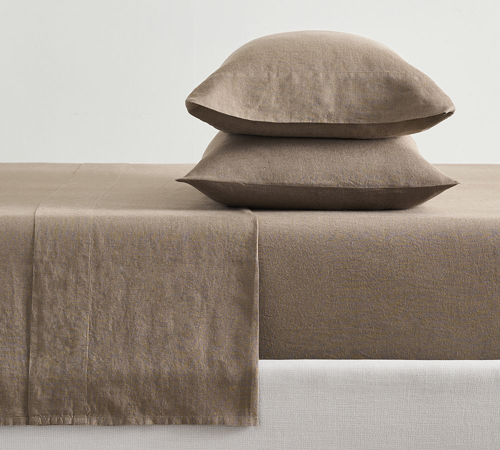 A neatly made bed with a brown linen sheet set. Two pillows are stacked on top of each other, showcasing matching pillowcases. The bedframe appears to be white and the walls are plain, emphasizing the minimalist style.