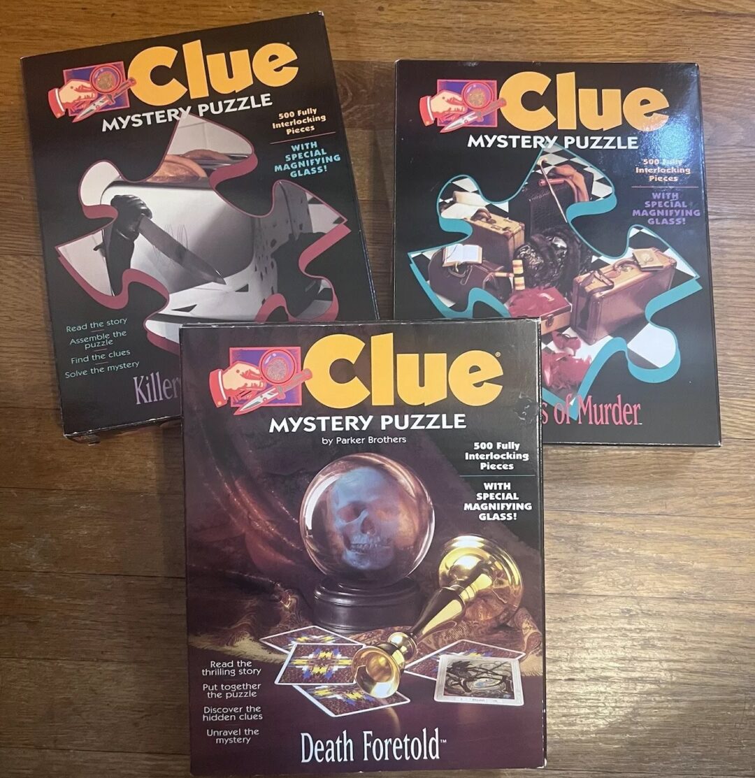 Three Clue mystery puzzle boxes are displayed on a wooden surface. Each box features different artwork with titles: "Killer Pieces," "The Sands of Murder," and "Death Foretold," depicting mysterious scenes.