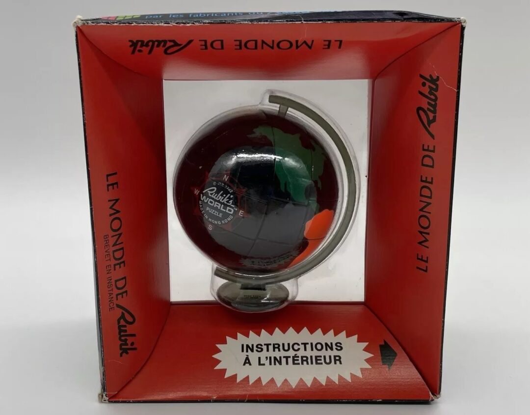 A boxed Rubik's World, featuring a globe in a red and black package. The box has "Le Monde de Rubik" and "Instructions à l'intérieur" written on it. The globe is mounted on a stand and encased within the box.