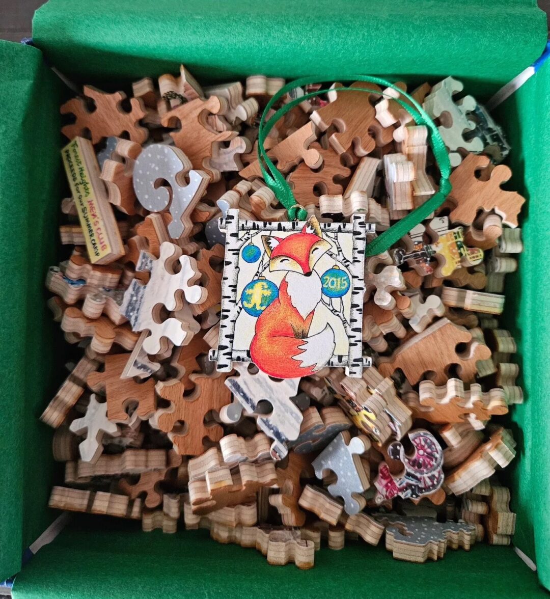 A box filled with wooden puzzle pieces featuring various shapes. On top is a completed piece depicting an illustrated fox surrounded by the year "2015." The puzzle piece has a green string attached, suggesting it could be used as an ornament.