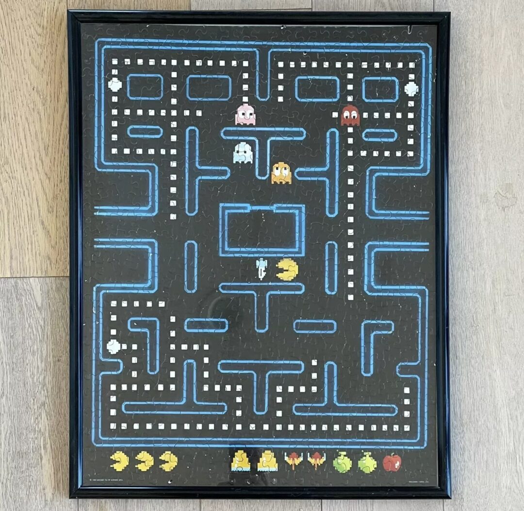 Framed artwork depicting a Pac-Man game maze with blue pathways on a black background. The scene includes Pac-Man, ghosts, and various fruits at the bottom.