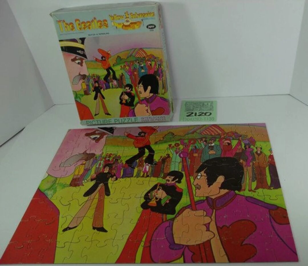 A partially completed puzzle is displayed in front of its box, featuring vibrant cartoon characters in various poses and colorful outfits. The puzzle box is labeled "The Beatles Yellow Submarine" with similar artwork.