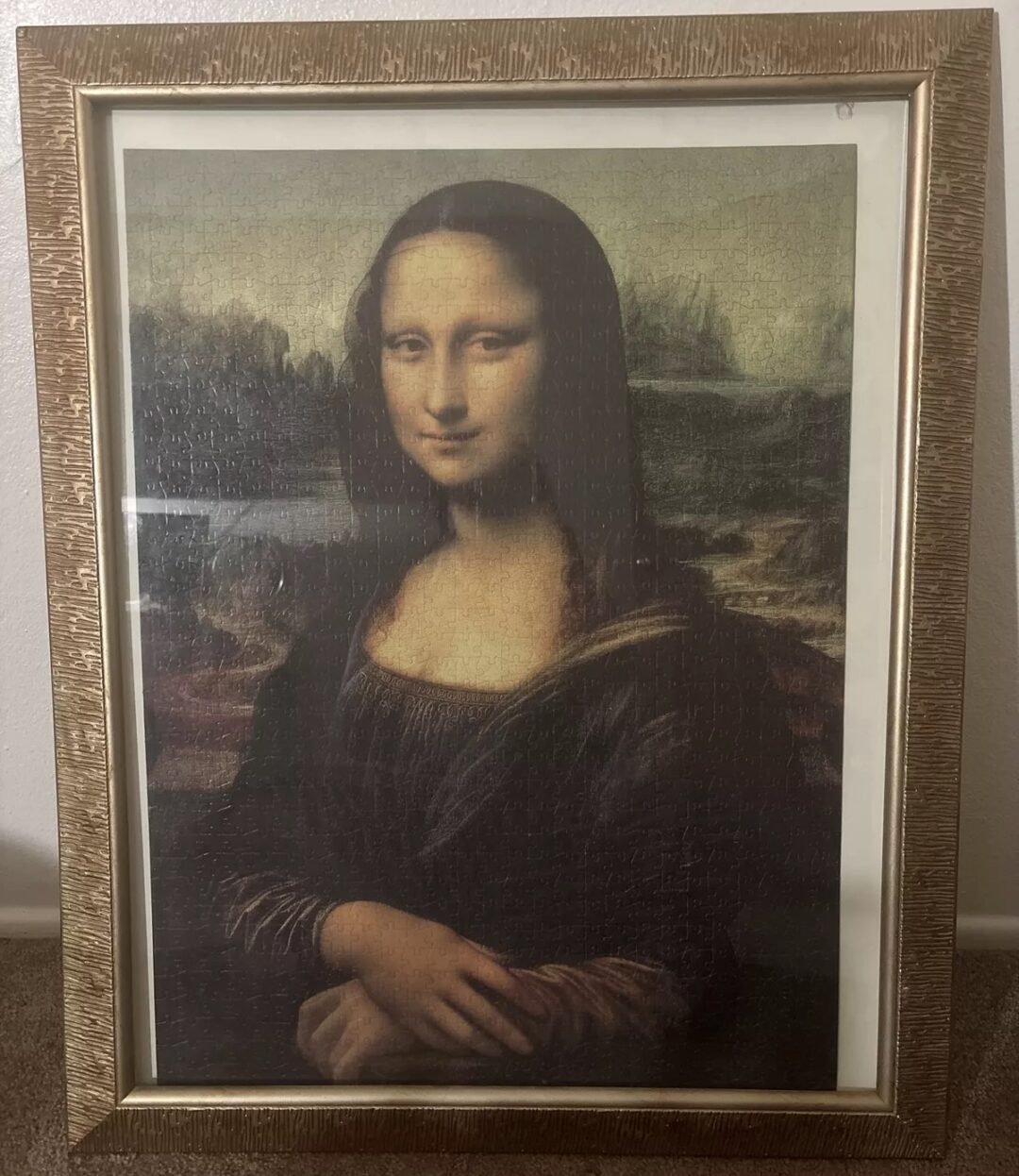 A framed replica of the Mona Lisa painting, set against a wall. The artwork features the iconic half-length portrait of a woman with a serene expression, sitting against a landscape background. The frame is textured and gold-colored.