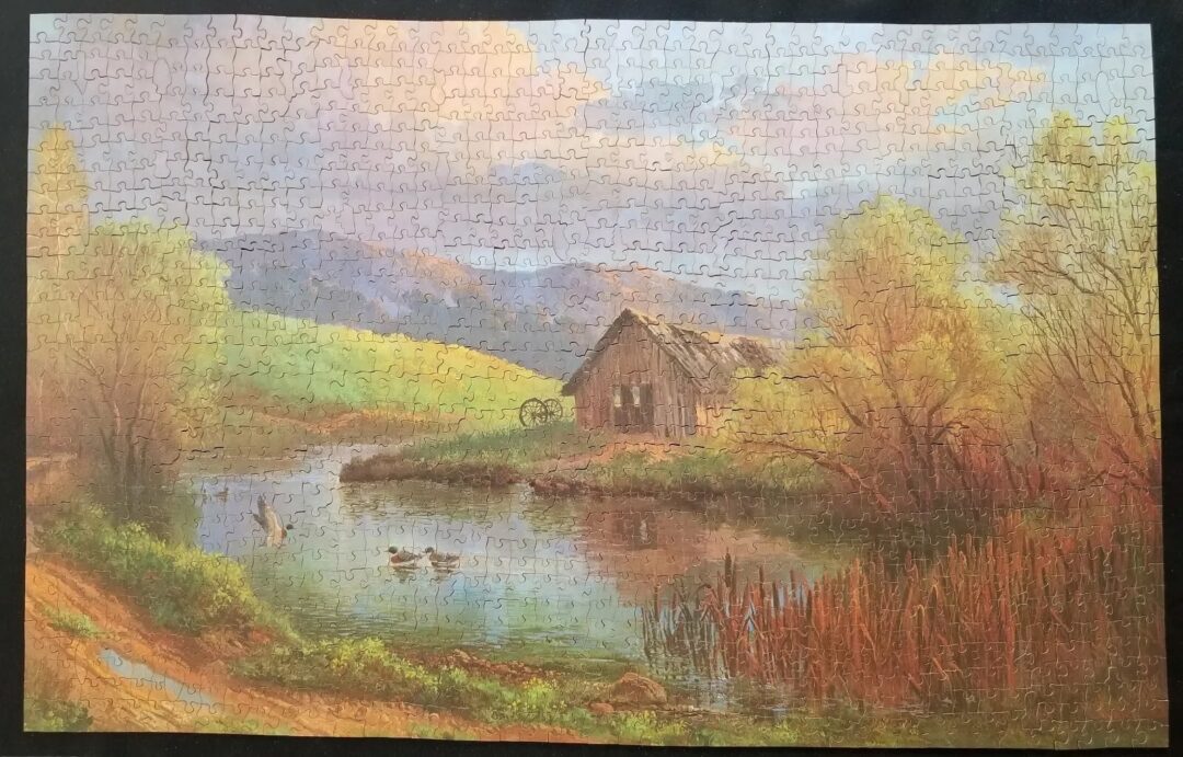 A completed puzzle depicting a serene landscape with a rustic wooden cabin by a reflective pond, surrounded by lush greenery and trees. A few ducks swim in the water under a sky with pink and blue clouds.