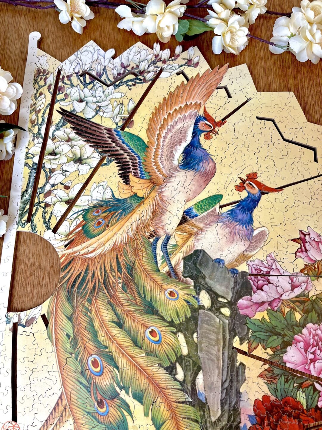 A partially completed jigsaw puzzle depicting two colorful peacocks perched on a tree with lush plumage. The background features blooming flowers and branches. Near the puzzle, there are additional white flowers and a fan-shaped layout.