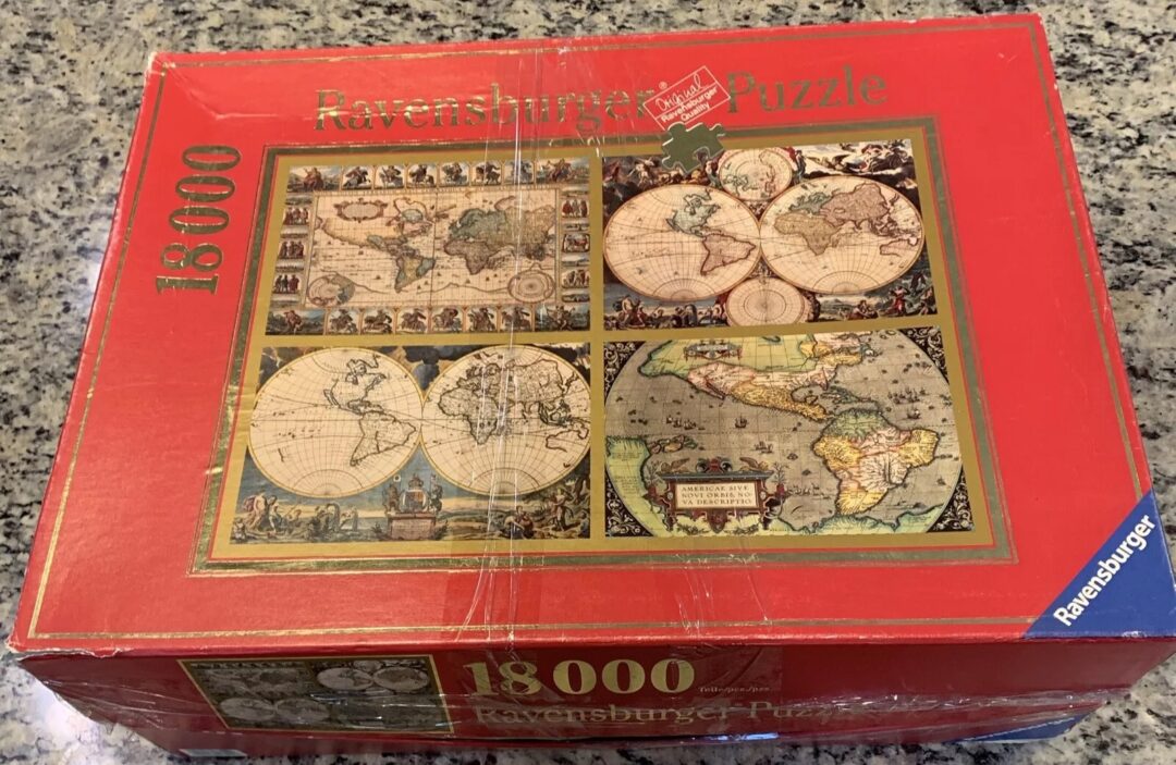 A Ravensburger puzzle box with a red background sits on a granite countertop. The box features an 18,000-piece puzzle depicting vintage maps with a world map illustration on the front.