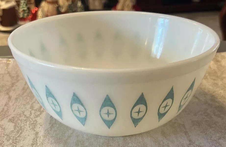 A vintage white bowl with a pattern of blue teardrop shapes, each containing a cross, is placed on a light-colored surface. The background shows blurred, decorative objects.