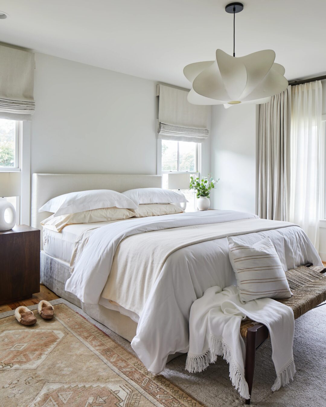 A serene bedroom with a large white bed, layered with pillows and a throw. A wooden bench at the foot holds a blanket. A modern, white ceiling light hangs overhead. Natural light filters through sheer curtains, highlighting a neutral area rug and slippers.