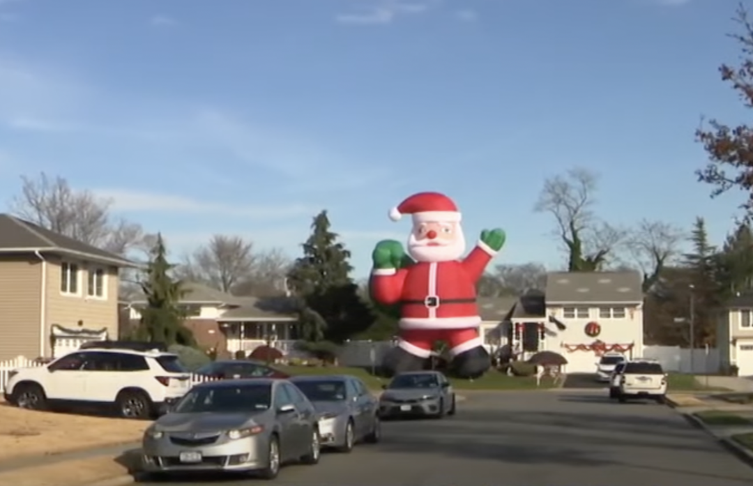 A giant inflatable Santa Claus stands on a residential street. Several cars are parked along the road, and houses with Christmas decorations are visible under a clear blue sky.