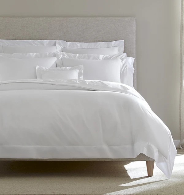 A neatly made bed with white linens and multiple pillows, set against a light gray upholstered headboard. Soft natural light filters through a window on the right, creating a calm and inviting atmosphere in the bedroom.