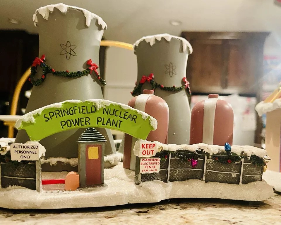 A decorative model of the Springfield Nuclear Power Plant, featuring cooling towers and buildings adorned with snow and wreaths. Signs read "Authorized Personnel Only" and "Keep Out." Small details include fences and snow-covered roofs.