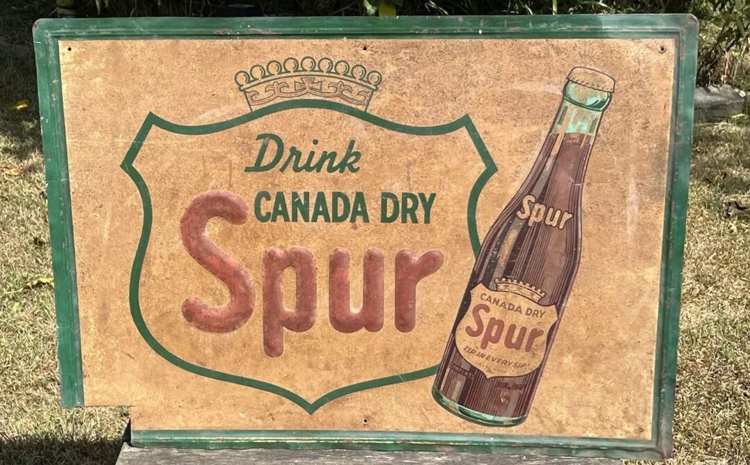 A vintage advertisement sign features a bottle of Canada Dry Spur with the words "Drink Canada Dry Spur" in bold letters. The sign has a rustic background and is placed on a grassy surface.