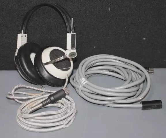 A pair of over-ear headphones with black and white design is placed on a gray surface. Next to them are two coiled cables, one shorter with an attached connector and one longer, both with similar design and color.