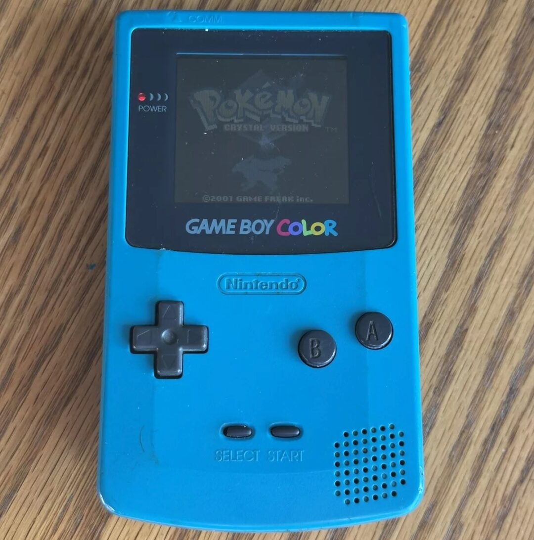 A blue Game Boy Color on a wooden surface, displaying the start screen of a Pokémon game. The device features a D-pad, A and B buttons, and Start and Select buttons.