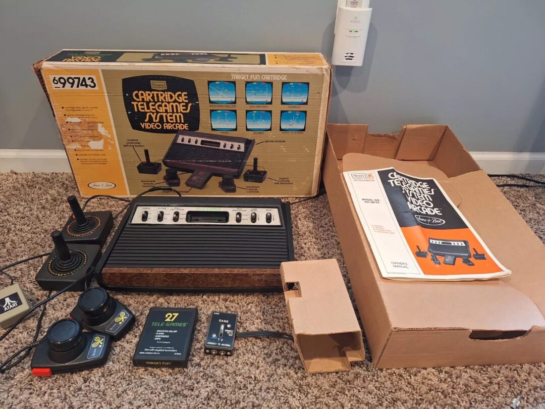 An old gaming console setup with a cartridge video game system in a box labeled "Cartridge Telegame," controllers, a catalog, and an adaptor, all placed on a carpeted floor.