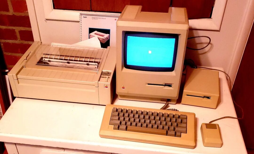 A vintage computer setup on a white desk, featuring an old beige monitor with a blue screen, a beige keyboard, a mouse, a floppy disk drive, and a dot matrix printer. A brick wall and a wooden cabinet are in the background.