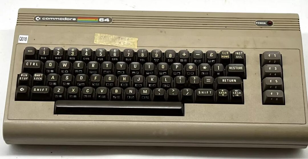 A vintage Commodore 64 computer with a beige casing and a black keyboard featuring various keys, including a large "RETURN" key. The top left corner has a logo, and there's a "POWER" indicator on the right.