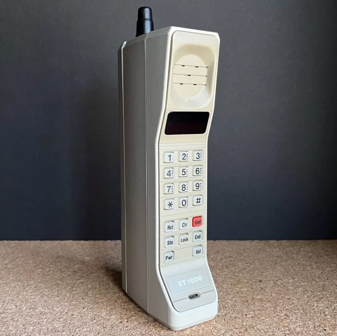 Vintage beige mobile phone with an antenna and numeric keypad, standing on a textured surface against a dark background. The phone features a small screen and buttons, marked with "ET 1000" at the bottom.