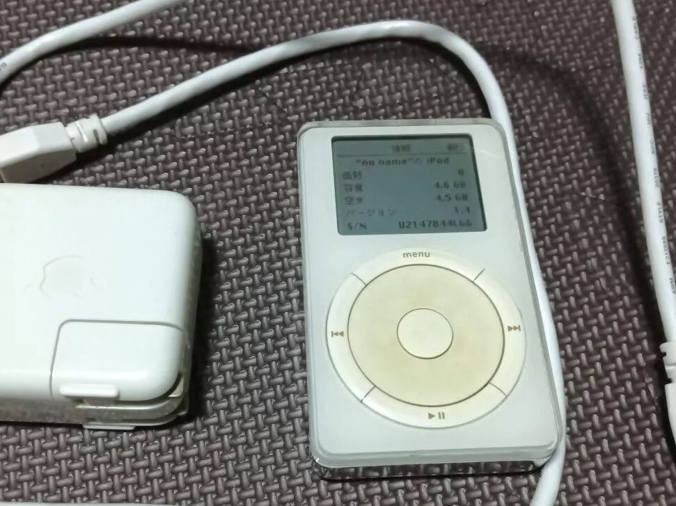 A vintage iPod with a small screen and circular control wheel is placed on a textured surface. It is connected to a charger with a white cable and power adapter featuring the Apple logo.