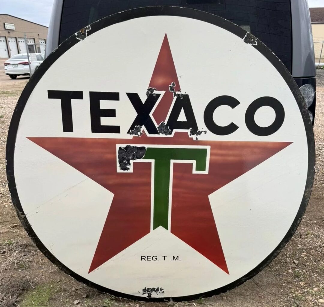 A large, round vintage Texaco sign featuring a red star with a green "T" in the center. The text "TEXACO" is displayed across the top, with "REG. T. M." below. The sign stands outdoors on a gravelly surface with buildings in the background.