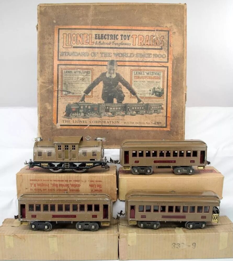 Vintage Lionel electric toy train set with a locomotive and three passenger cars in front of an original box. The box displays classic Lionel branding and graphics, showcasing the historical style of the early 1900s.