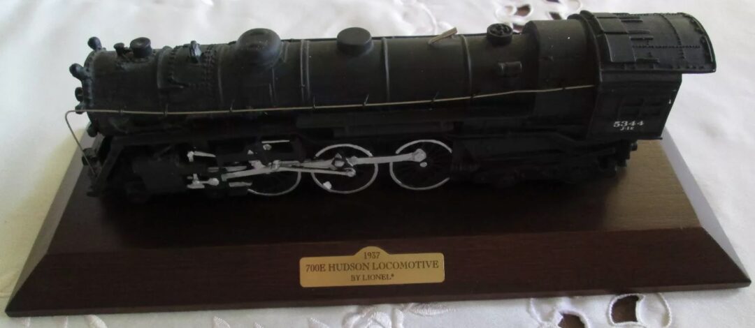 A detailed model of a black 1937 700E Hudson Locomotive by Lionel is displayed on a wooden base with a gold plaque. The locomotive features intricate wheel and rail details, sitting on a white lace tablecloth.