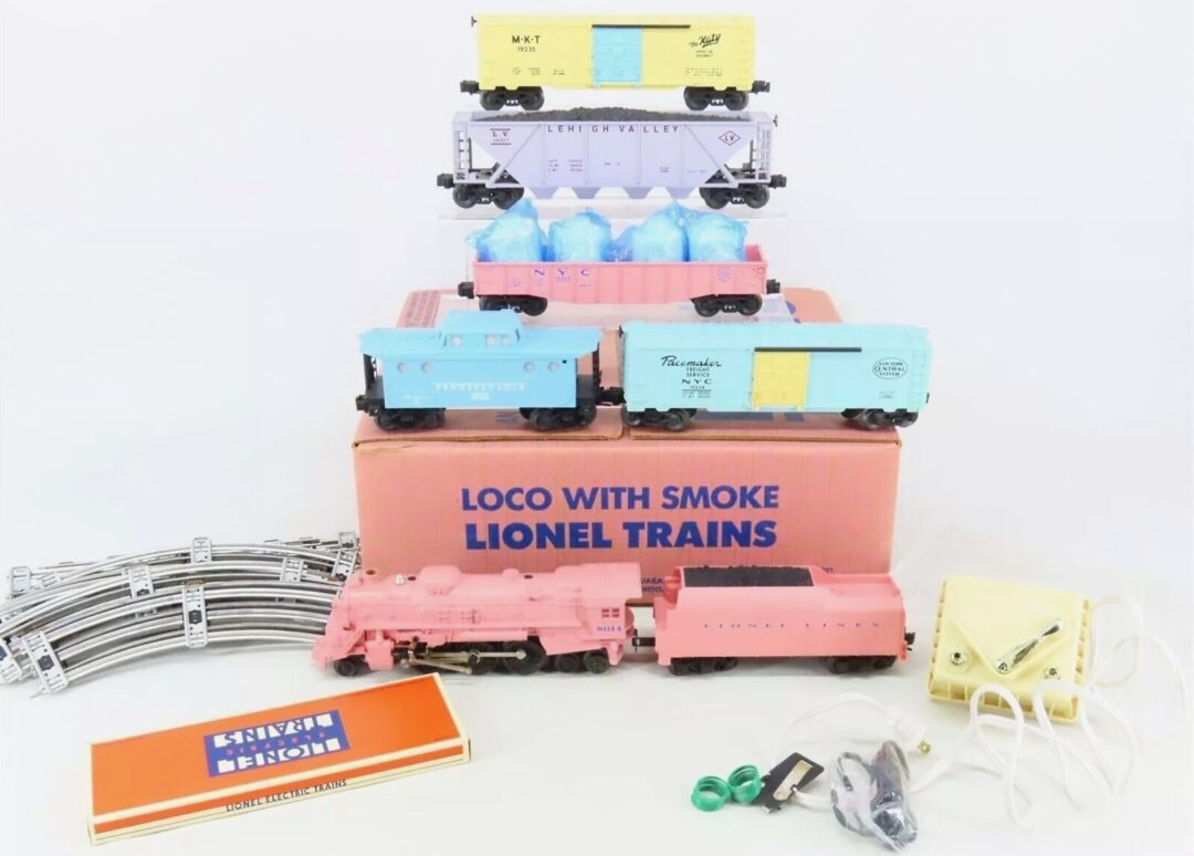 Vintage Lionel toy train set with tracks, a pink locomotive, multiple colorful boxcars, and accessories on display. Items are arranged around an orange box labeled "Loco with Smoke Lionel Trains.