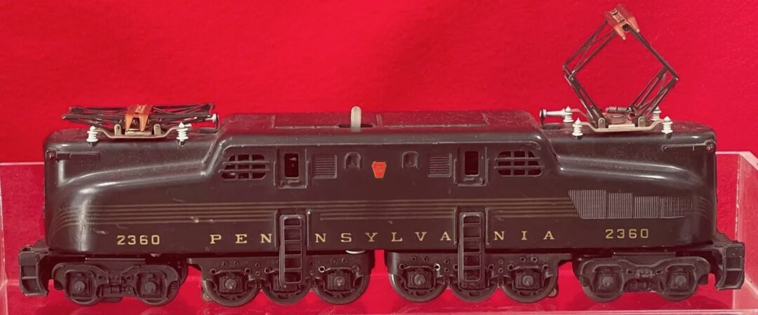 A model of a Pennsylvania Railroad locomotive numbered 2360, set against a red background. The model features intricate detailing with pantographs raised on top.