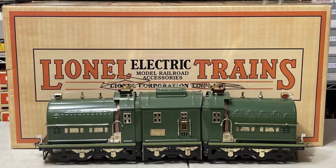 A green model electric train is displayed in front of a large orange and beige box labeled "Lionel Electric Trains." The train features detailed designs and the text "Lionel Lines" on its side.