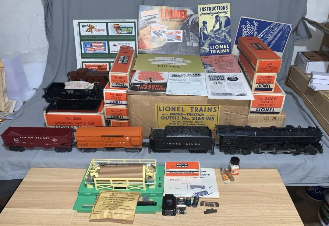 A collection of vintage Lionel train sets, including various train cars, a locomotive, instruction booklets, and original packaging, is displayed on a wooden table and a folded grey cloth backdrop.