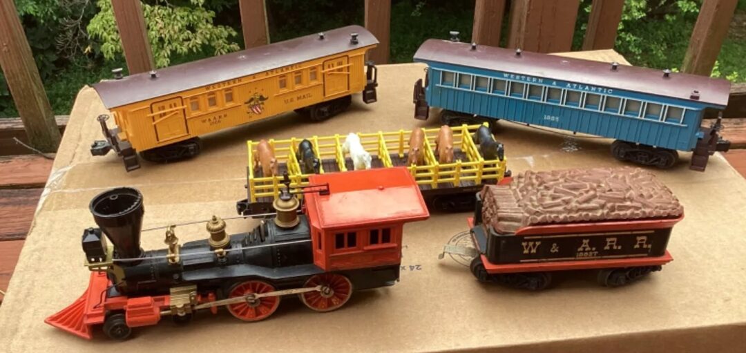 Vintage model trains displayed on a cardboard box. The set includes a red and black locomotive with a coal tender, a yellow livestock car, a blue passenger car, and a yellow freight car. Wooden railing and greenery are visible in the background.