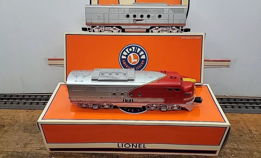 A model train locomotive in red and silver sits atop an orange and white Lionel box. Behind it, the box showcases an illustration of the same locomotive model in gray. A round Lionel emblem is visible above.