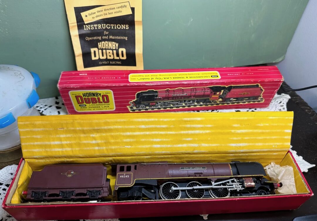 A vintage Hornby Dublo train set is displayed, including a locomotive and tender in their original box. An instruction manual is partially visible, and a decorated tabletop with lace coasters surrounds the set.